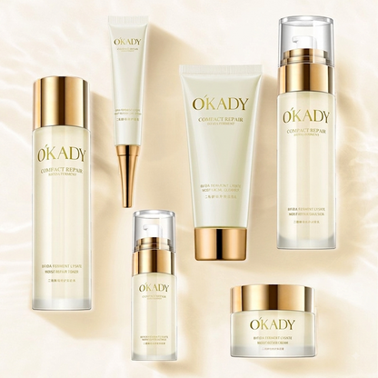Six OKADY skincare products arranged on a soft cream background: two tall bottles, two tubes, one pump bottle, one small pump bottle, and one jar. The products are frosted off-white with metallic gold caps and gold branding.